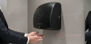 Homem de terno estendendo as mãos sob um secador de mãos automático preto instalado na parede de um banheiro moderno com azulejos claros. Equipamento sem contato, moderno e sustentável, refletido no espelho ao lado.